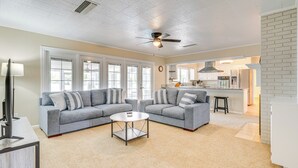 A spacious living room with two grey sofas, a round coffee table, and a ceiling fan. A kitchen is visible in the background with white cabinets and a bar counter.
