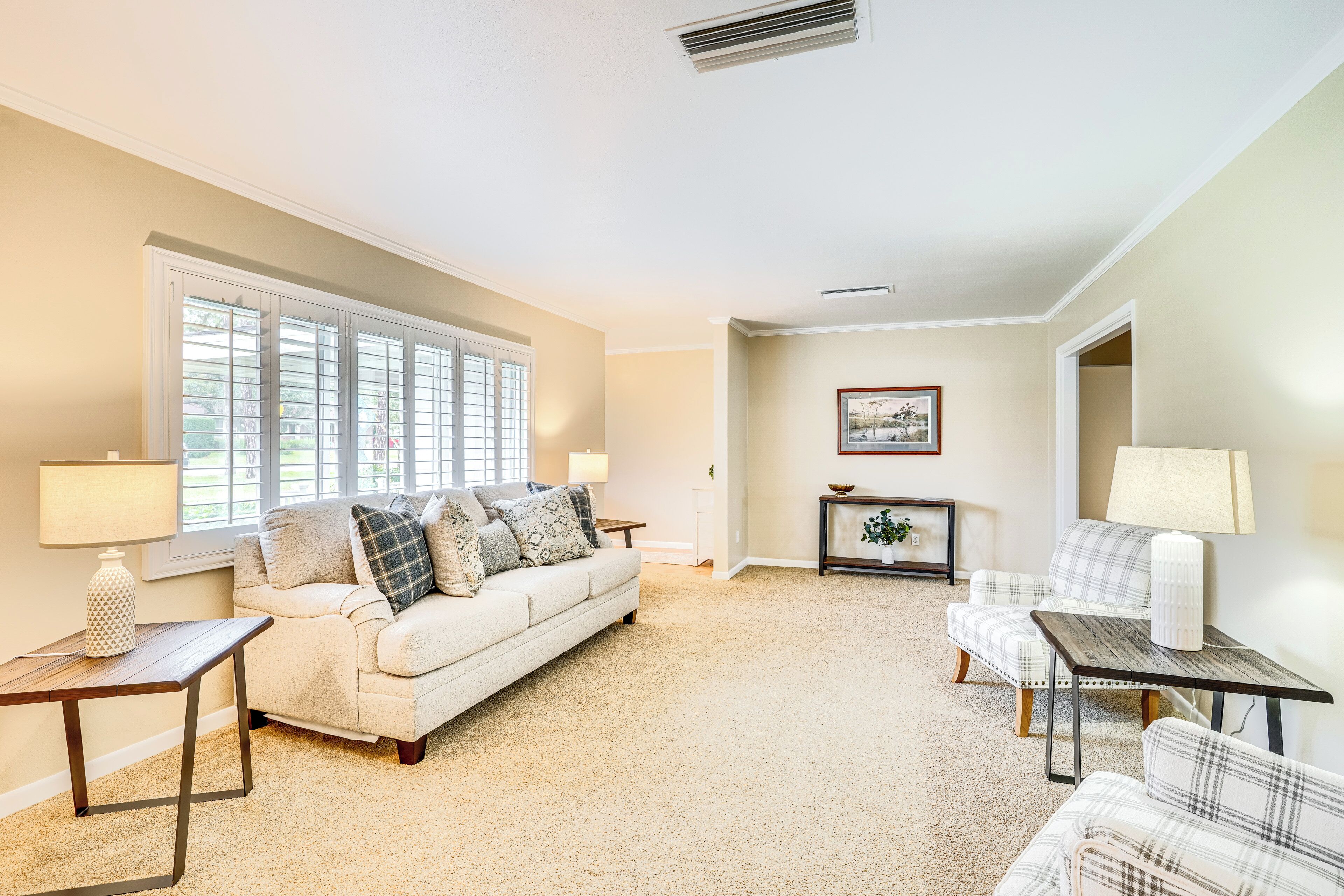 A living room with a beige carpet, a white sofa with pillows, a plaid armchair, a small table with a lamp, and a window with blinds.