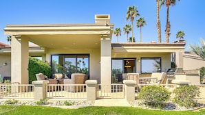 A covered outdoor patio with wicker furniture, a stone pathway, and a view of palm trees. - Community Pools & Golf Views: Palm Desert Condo! (Palm Desert)