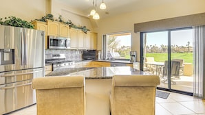 A modern kitchen with stainless steel appliances, a large island with bar stools, and a view of a golf course through the sliding glass doors. - Community Pools & Golf Views: Palm Desert Condo! (Palm Desert)