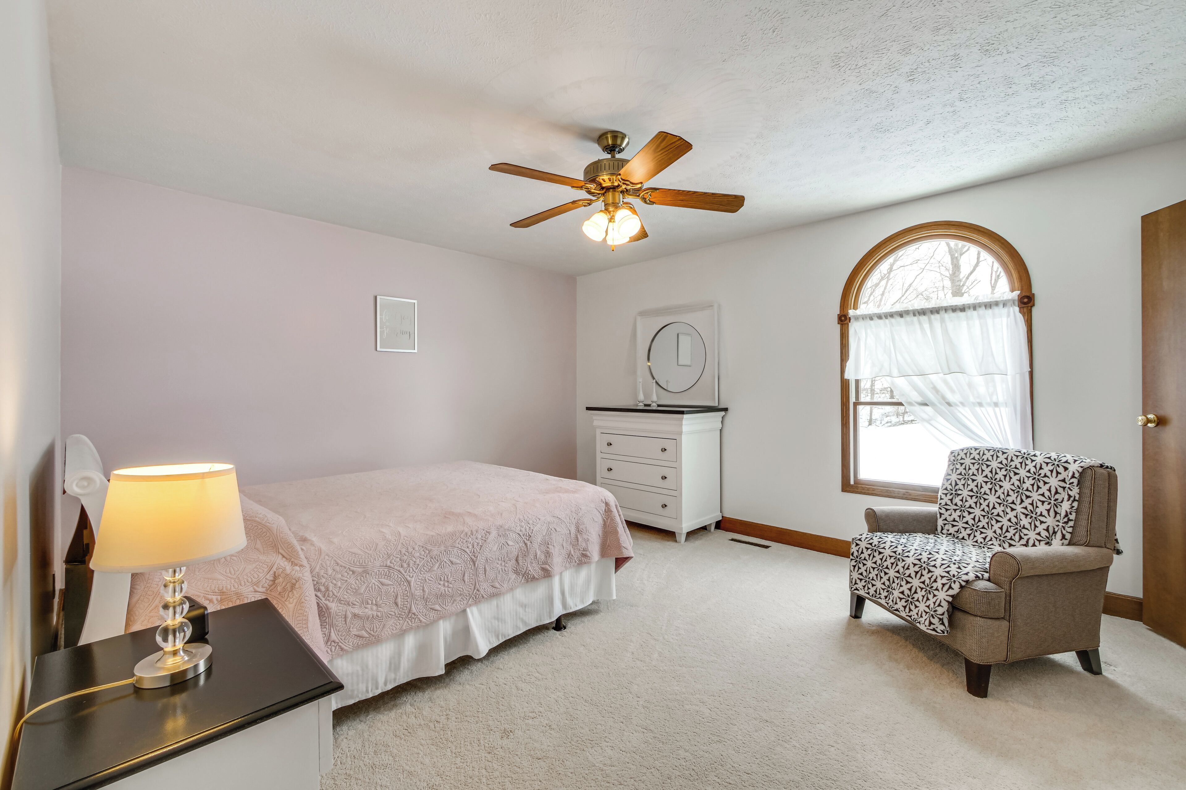 Une chambre à coucher comprenant un lit, une chaise, une table de chevet, une commode, un ventilateur de plafond, une fenêtre avec des rideaux et une porte.