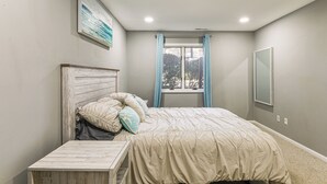 Una camera da letto con un letto, un comodino, una finestra con tende, uno specchio fissato alla parete e un quadro appeso al muro.