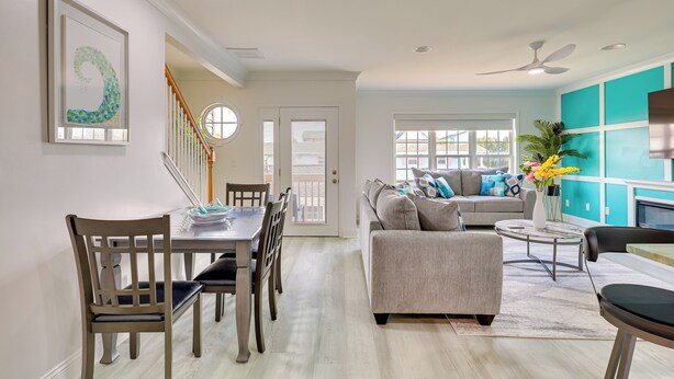 A modern living room with a grey sofa, a dining table with chairs, and a staircase. - Walk to Ocean: Carolina Beach Condo w/ 2 Balconies (Carolina Beach)