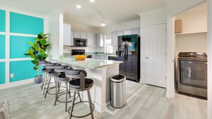 A modern kitchen with white cabinets, a central island with bar stools, a stainless steel refrigerator, and a built-in oven. - Walk to Ocean: Carolina Beach Condo w/ 2 Balconies (Carolina Beach)