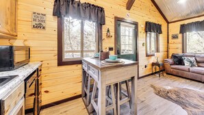 A cozy cabin interior with wooden walls, a kitchen area with a stove, a dining table, and a couch. - 'bluebird Treehouse' Near Kiamichi Mtns w/ Hot Tub (Talihina)