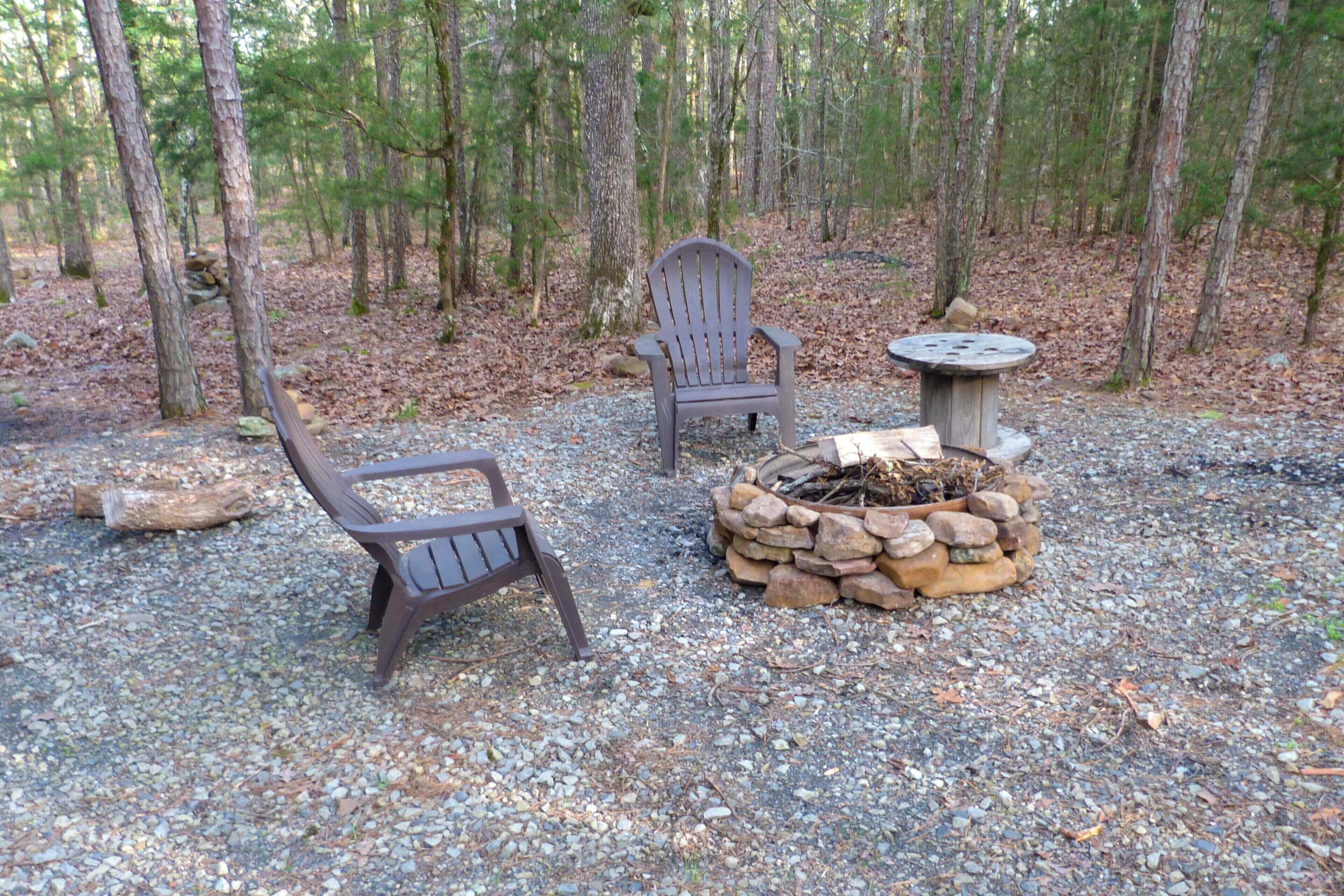 A forested area with two Adirondack chairs and a stone fire pit.