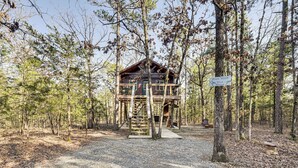 A treehouse with a wooden staircase leading up to a deck, surrounded by a forest. - 'bluebird Treehouse' Near Kiamichi Mtns w/ Hot Tub (Talihina)