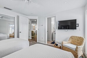 Habitación de hotel con dos camas, televisión, una silla de mimbre y un pasillo que conduce a un comedor.