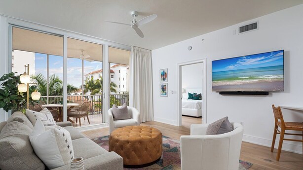 Un salon spacieux doté d’une grande fenêtre, d’un ventilateur de plafond, d’un téléviseur à écran plat et d’un balcon aménagé avec du mobilier d’extérieur.
