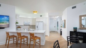 Une cuisine moderne avec des meubles blancs, un îlot central doté de tabourets de bar, un coin repas avec une table et des chaises noires, et un salon avec un téléviseur et des œuvres d’art.
