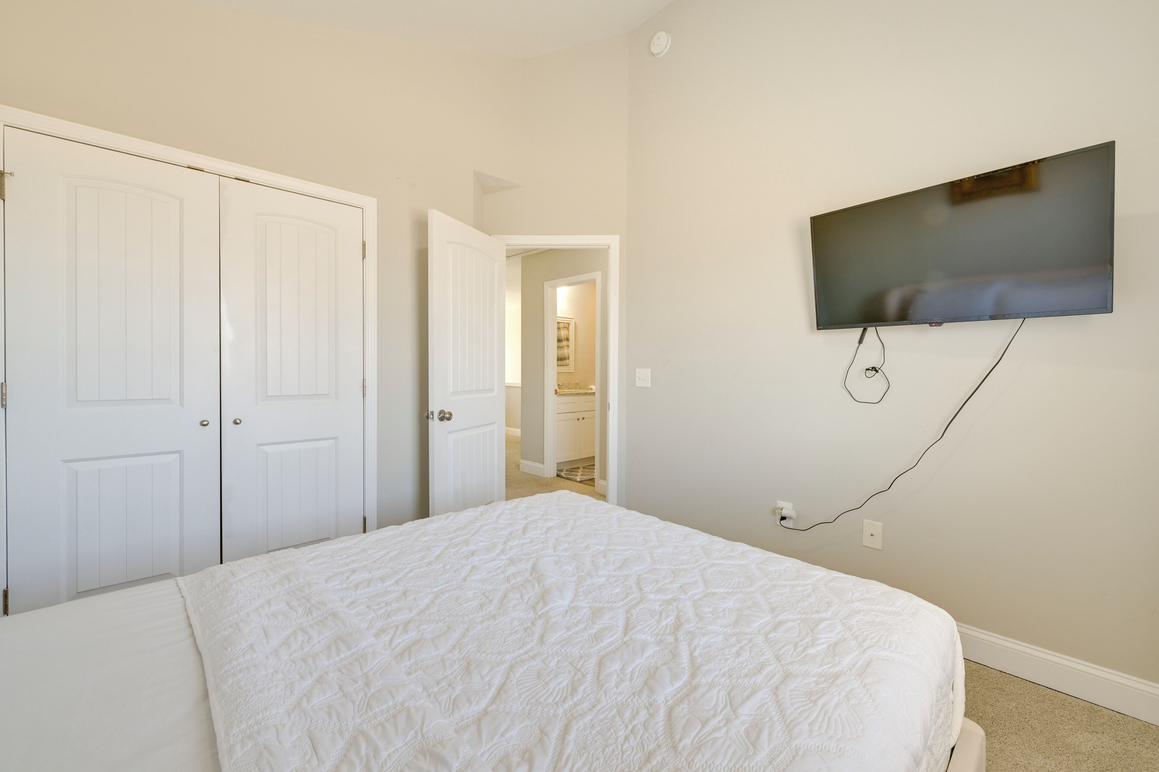 Une chambre à coucher avec un grand lit, un téléviseur fixé au mur et une porte donnant sur une autre pièce.
