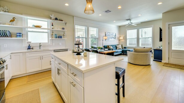 A modern kitchen with white cabinetry, a central island, and a dining area with a blue sofa and coffee table. - 1 Mi to Beach: Home w/ Pool Access in Port Aransas (Port Aransas)