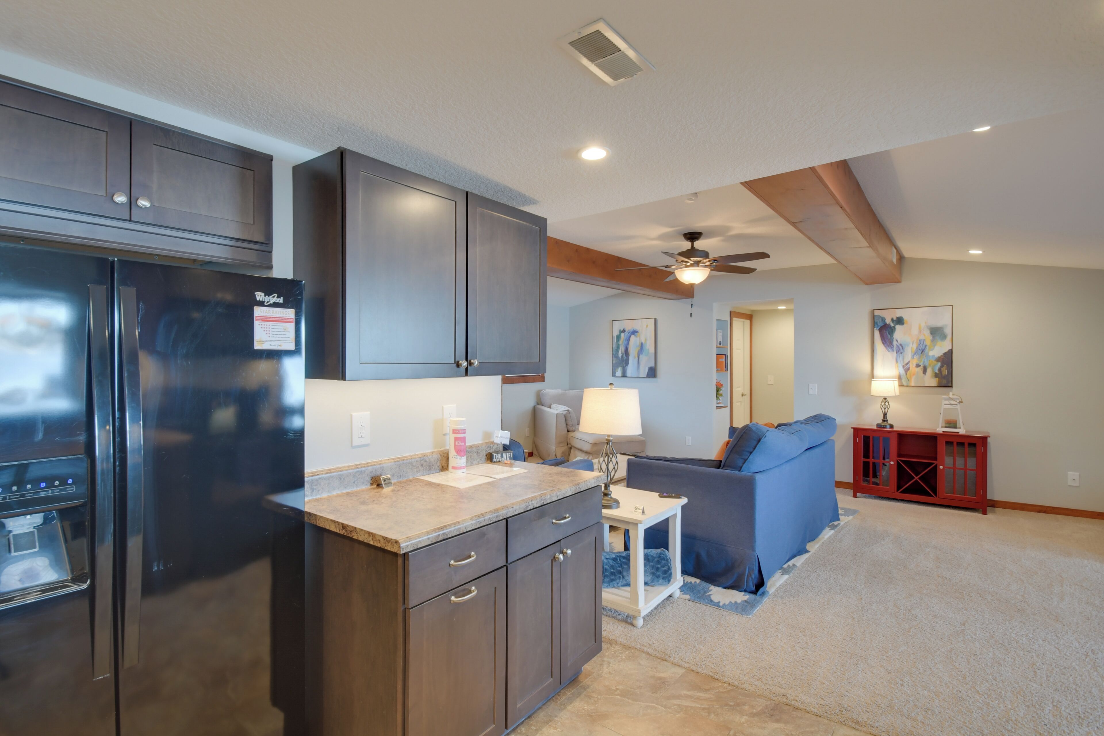 A modern kitchen with dark cabinetry, a stainless steel refrigerator, and a small island with a sink. There is a ceiling fan and recessed lighting.