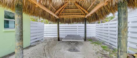 Un espacio al aire libre techado con una mesa y sillas, rodeado por una valla blanca y un muro verde.