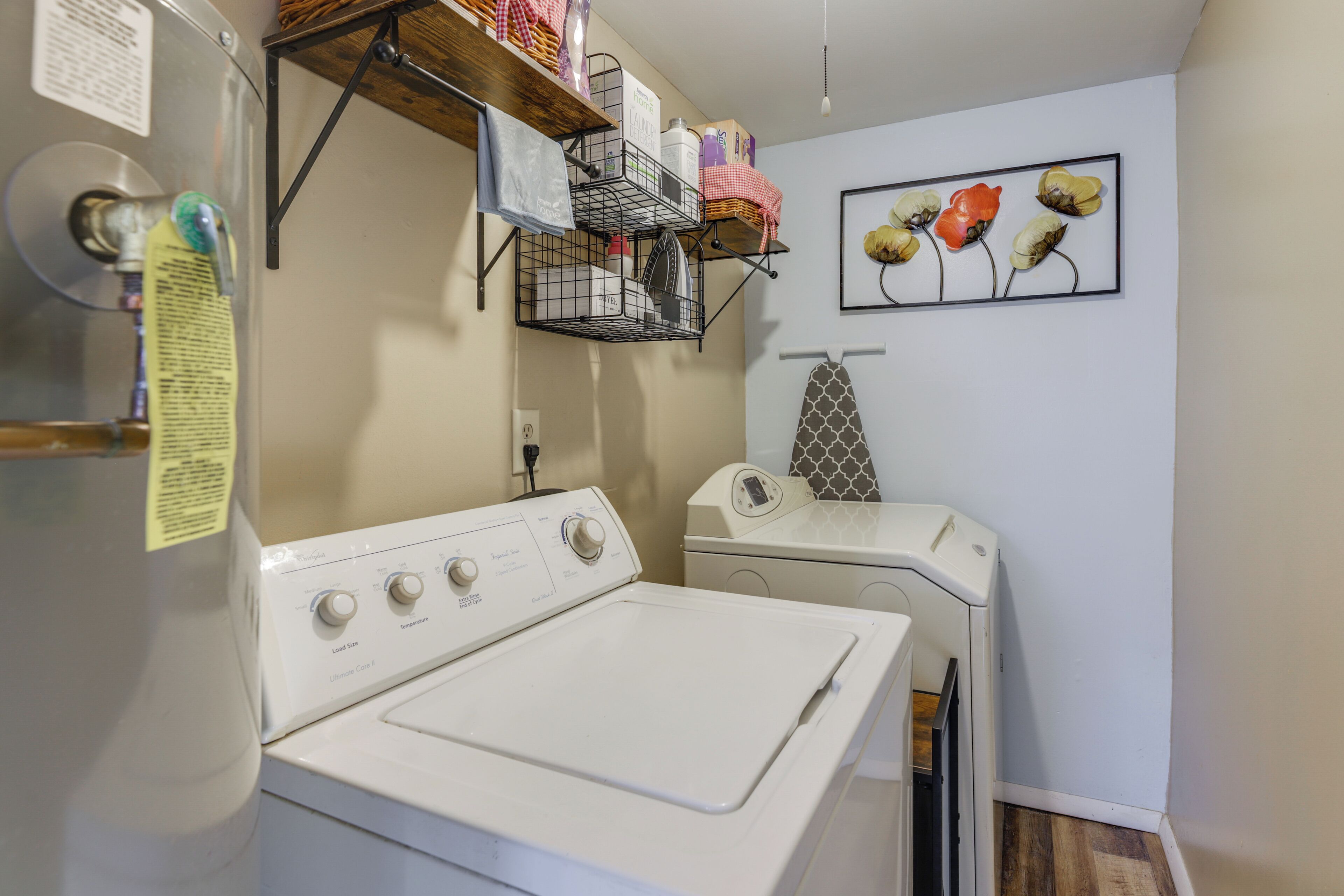 A laundry area with a front-loading washer and dryer, shelves above holding various items, and a wall-mounted ironing board.