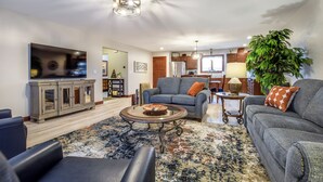 A spacious living room with a large flat-screen TV, a gray sofa, a coffee table, and a dining area with wooden cabinets and a chandelier. - 2026-NEW BOOKING SPECIAL at The Belvedere just Steps to downtown. (New Glarus)