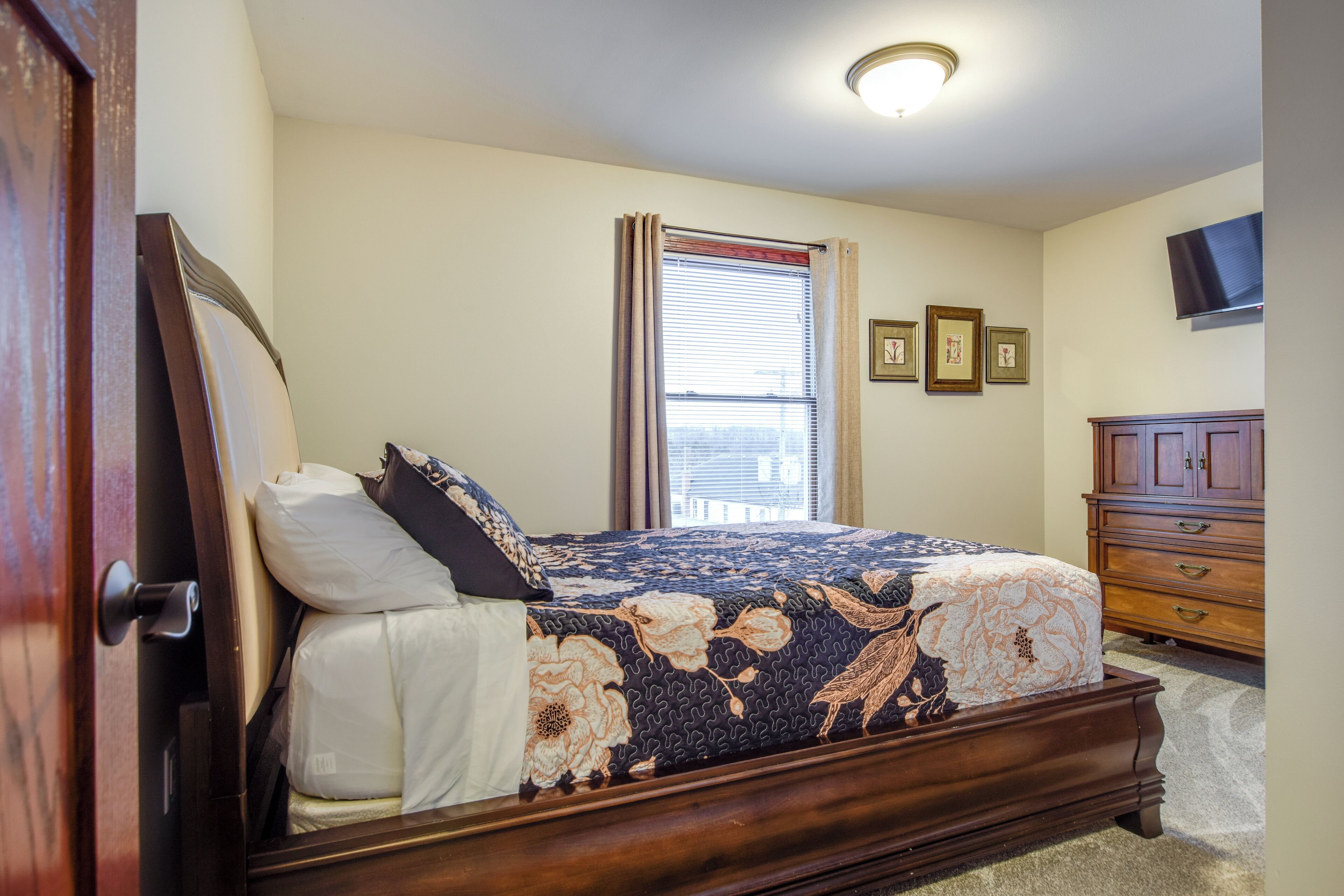 A bedroom with a bed, a window with blinds, a dresser, and a flat-screen TV mounted on the wall.