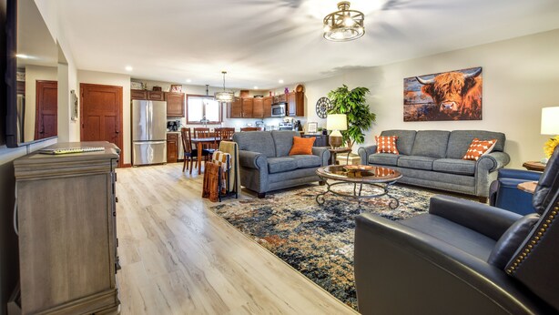 A spacious living room with a grey sofa, a black leather chair, and a coffee table. There is a kitchen in the background with wooden cabinets and a refrigerator. - 2026-NEW BOOKING SPECIAL at The Belvedere just Steps to downtown. (New Glarus)