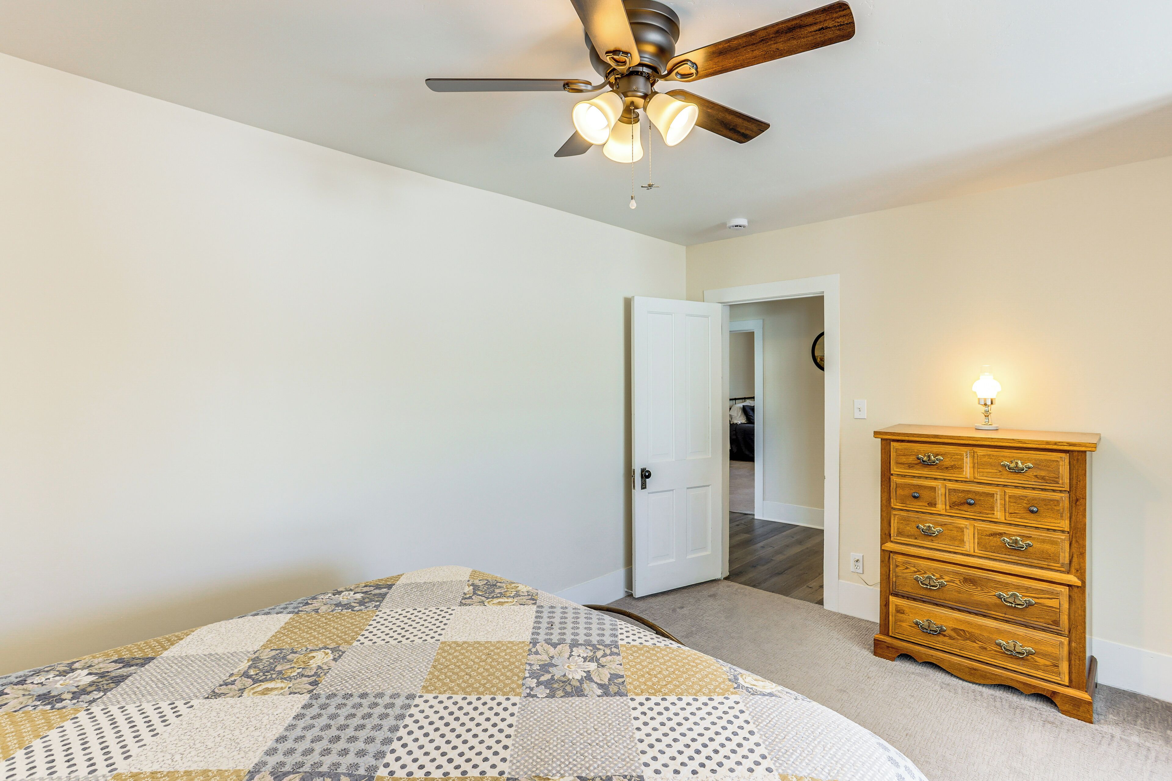 A bedroom with a bed, a wooden dresser, a ceiling fan, and a door leading to another room.