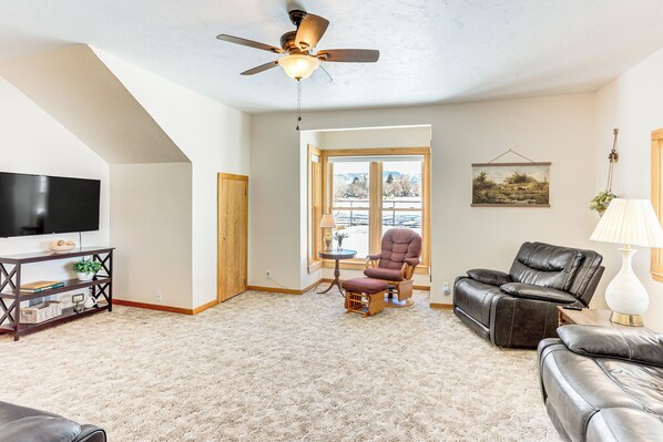 A living room with a ceiling fan, a flat-screen TV, a leather sofa, a recliner, and a large window offering a view of the outdoors. - Patio & Mountain Views: Farmhouse in Mancos! (Mancos)