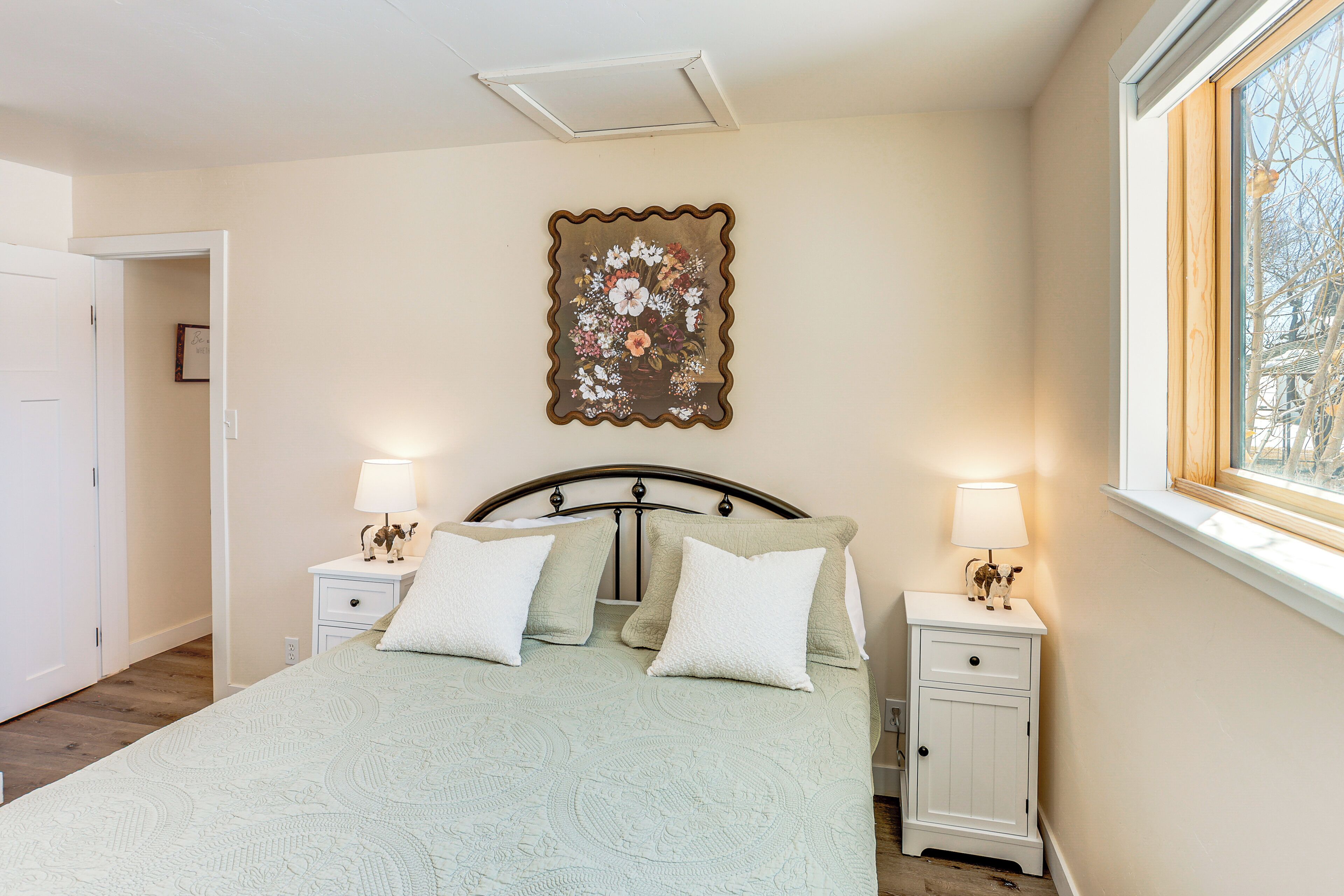 A bedroom with a bed, two bedside tables with lamps, a decorative wall piece, and a window with a view.