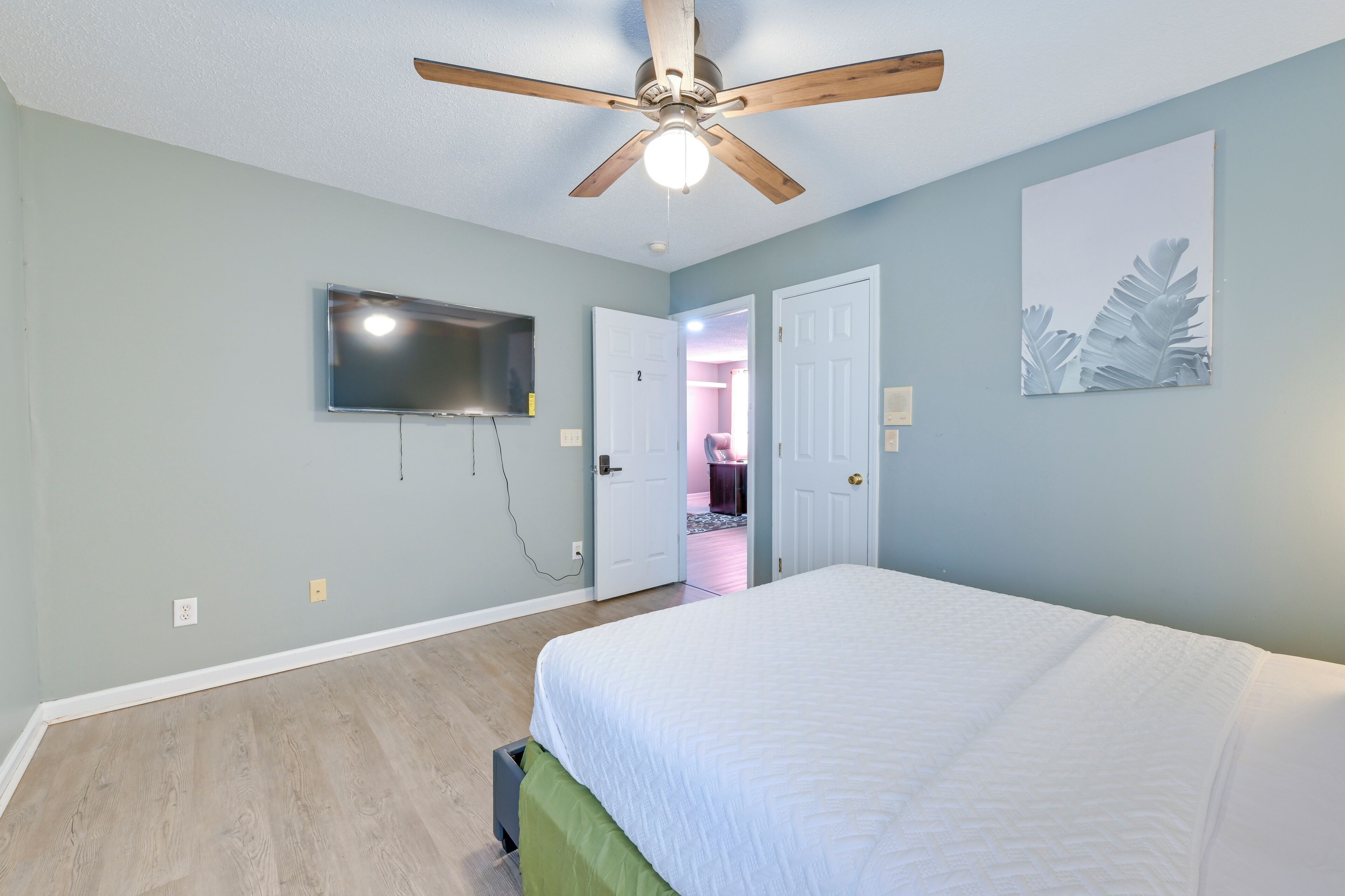 A bedroom with a bed, ceiling fan, TV, and artwork on the wall.