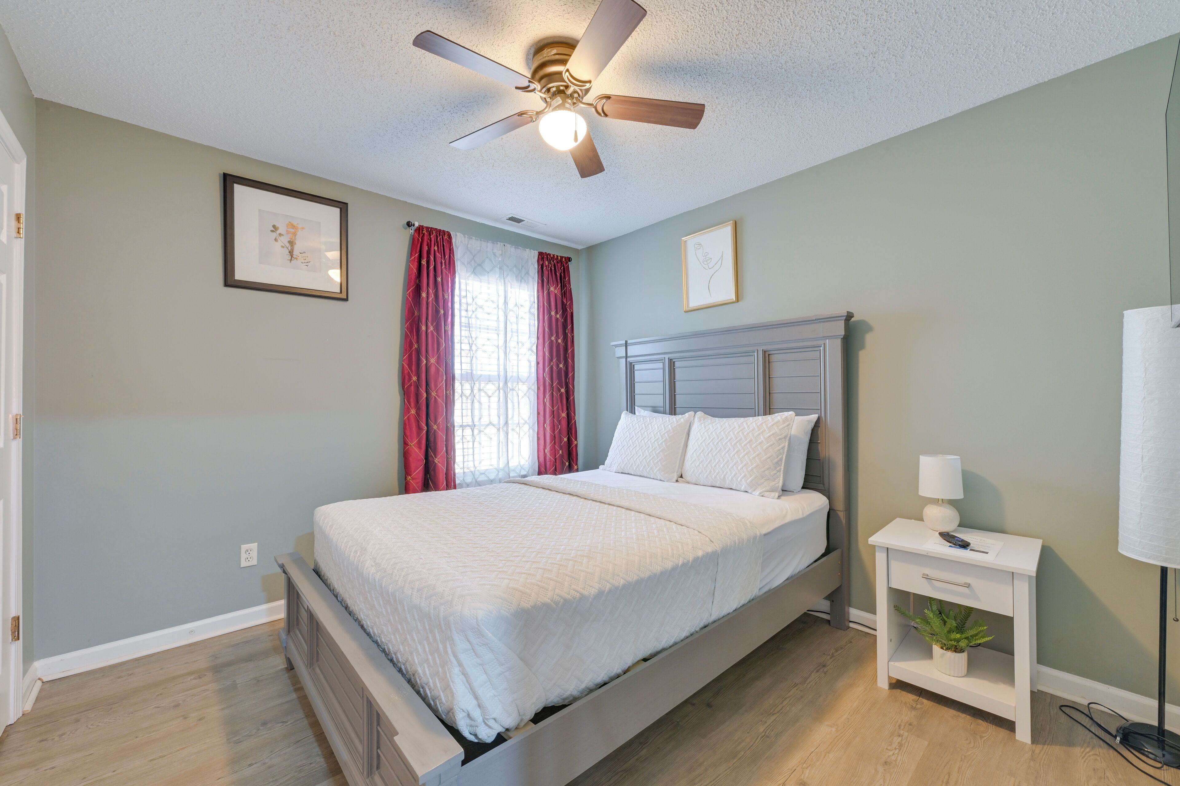 Une chambre à coucher avec un lit, une table de chevet, un ventilateur de plafond et une fenêtre avec des rideaux.