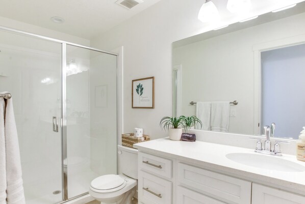 A modern bathroom with a glass shower enclosure, a white toilet, a vanity with drawers, and a large mirror. - Walk to Marina & Lake: Spacious Garden City Gem! (Garden City)