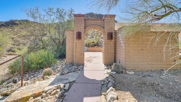 An arched brick entrance with a pathway leading through it, surrounded by desert vegetation and a rocky terrain.