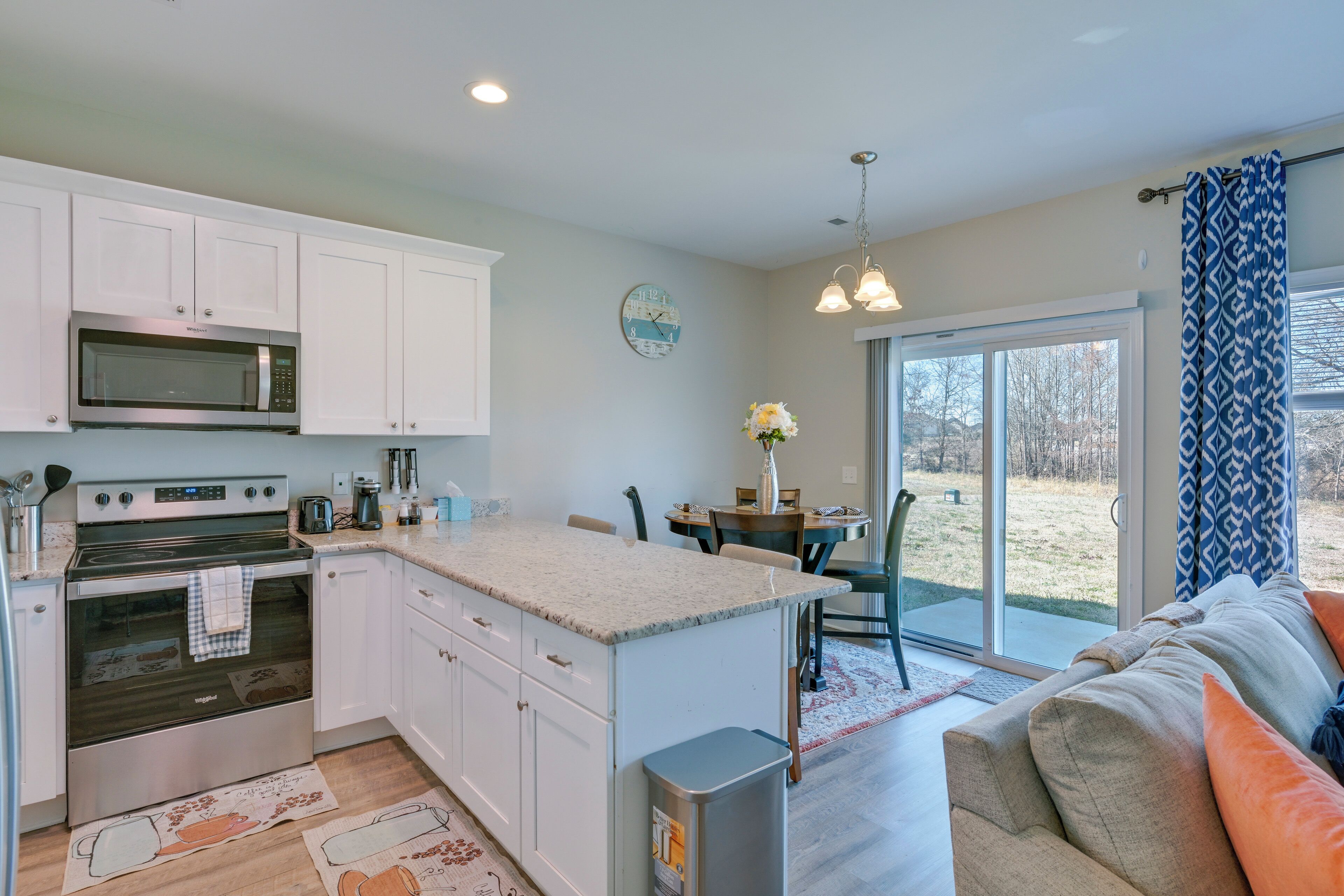 A modern kitchen with white cabinets, a stainless steel microwave, and an oven. There is a dining area with a table and chairs, and a large window with a view of the outdoors.