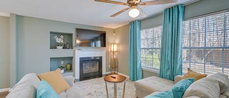 A living room with a fireplace, a ceiling fan, and a large window with curtains.