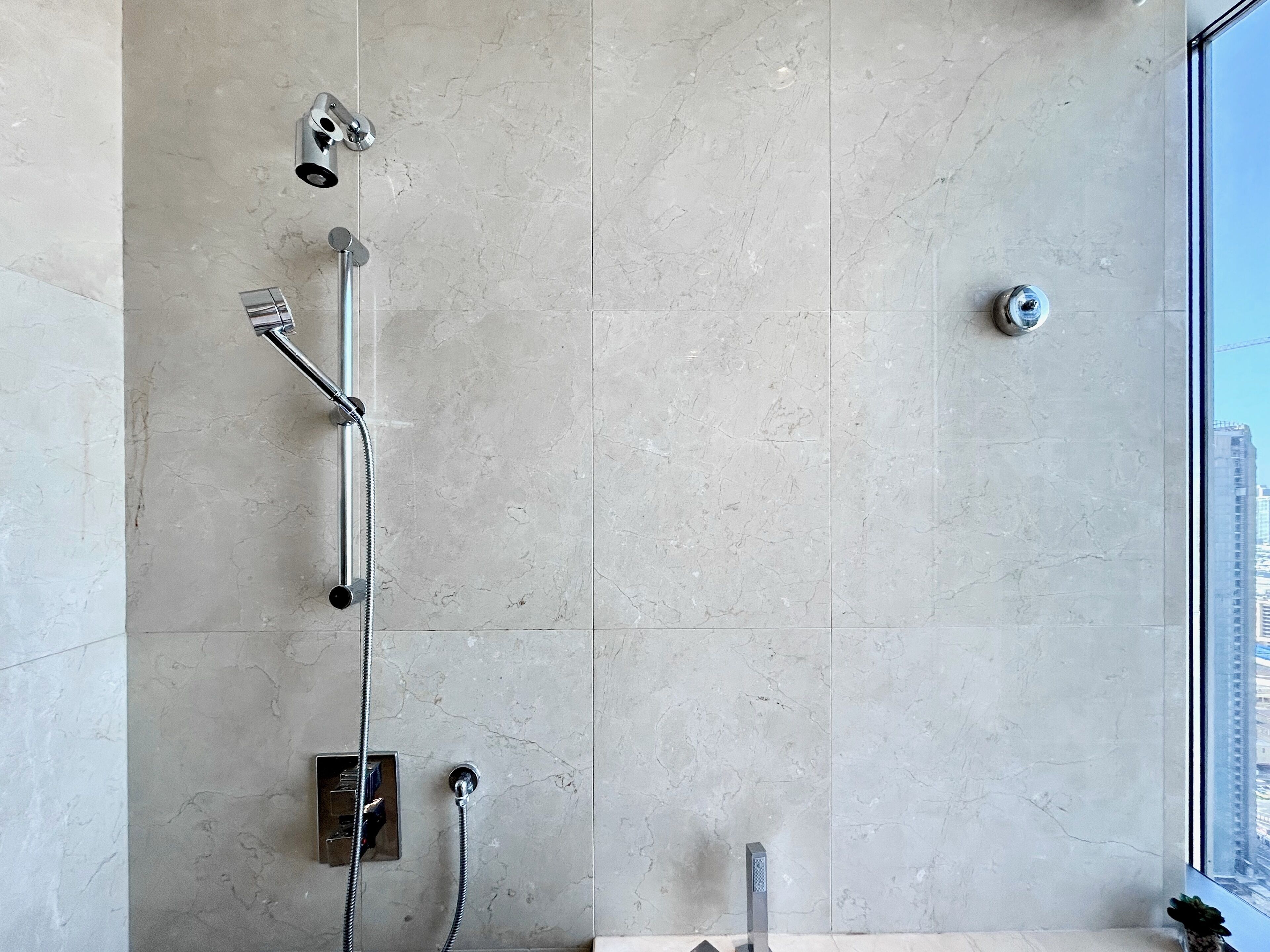 Un baño moderno con cabina de ducha de vidrio, panel de control de agua a la vista y vistas a un paisaje urbano a través de un amplio ventanal.