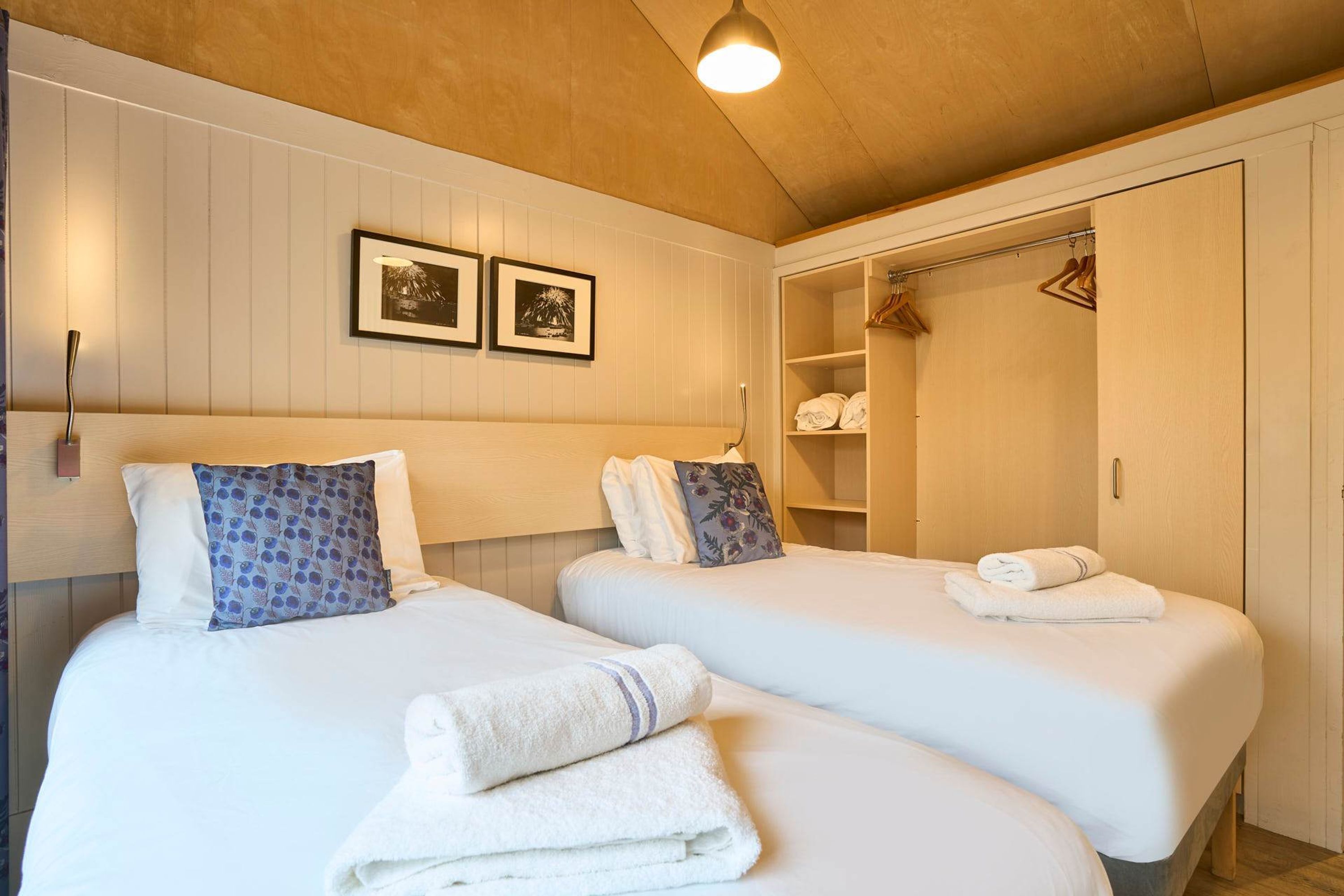 A hotel room with two beds, white bedding, a wooden ceiling, and a wardrobe with hanging clothes.