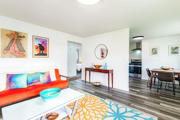 A modern living room with a red sofa, a coffee table, and a patterned rug. There is a dining area with a table and chairs, and a kitchen in the background.