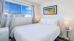 A hotel room with a large bed, a window with blinds, a framed picture on the wall, and a bedside table with a lamp.
