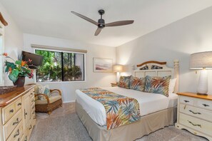 Un dormitorio con cama, ventilador de techo, mesita de noche, lámpara, cómoda, silla, ventana con vista a vegetación y televisor sobre un mueble.