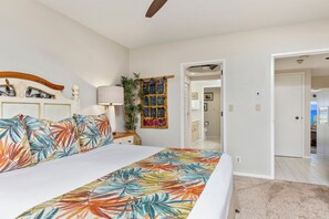 A bedroom with a bed, a nightstand, a lamp, a wall hanging, and a door leading to another room.