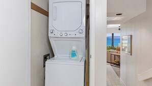 Un lavadero moderno y compacto, con lavadora y secadora integradas, una botella de detergente y vista al océano a través de una puerta abierta.