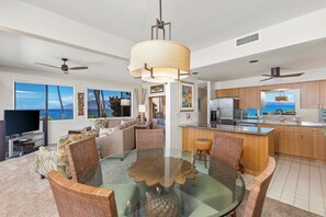 Un salon moderne avec une table à manger en verre, des chaises en osier, un téléviseur à écran plat et une vue sur l’océan.