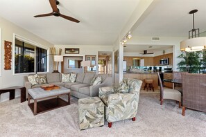 A spacious living room with a sofa, ottoman, and armchair, a kitchen in the background, and a ceiling fan.