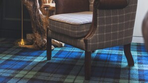 A tufted armchair with a plaid pattern, a wooden side table with a lamp, and a window with floral curtains. - Bein Inn hotel  (Perth)