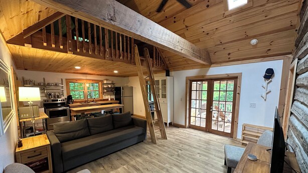 A cozy cabin interior with a wooden staircase, a kitchen area, a dining table, and a sofa. - Secluded Cabin on Beautiful Riverside Farm (State Road)