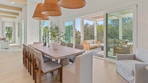 A spacious dining area with a long wooden table and chairs, surrounded by large windows offering a view of the outdoors. - Sleeps 20! Rooftop GULF VIEWS, Private Pool, LSV/golf cart, Bikes, 2 Kitchens! Pet Friendly! (Santa Rosa Beach)