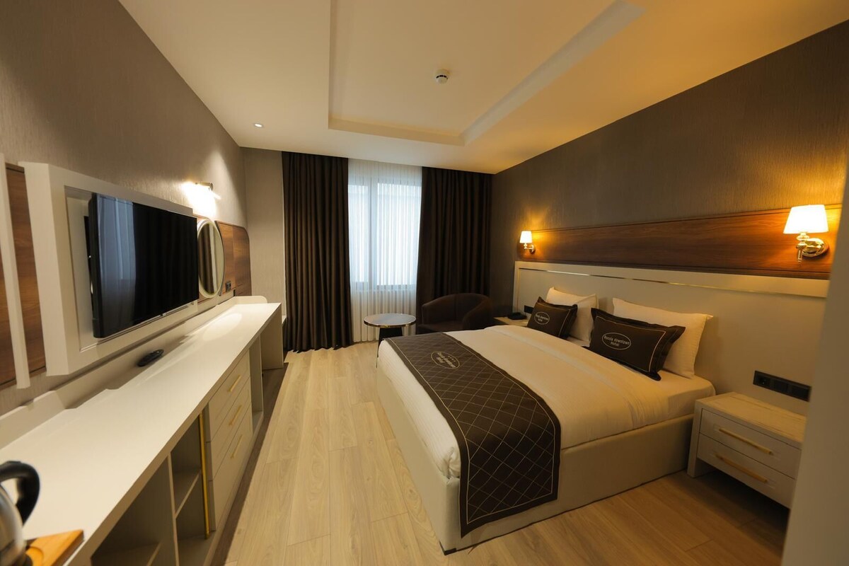 Quarto de hotel com cama, mesas de cabeceira, escrivaninha, cadeira, televisão e janela com cortinas.