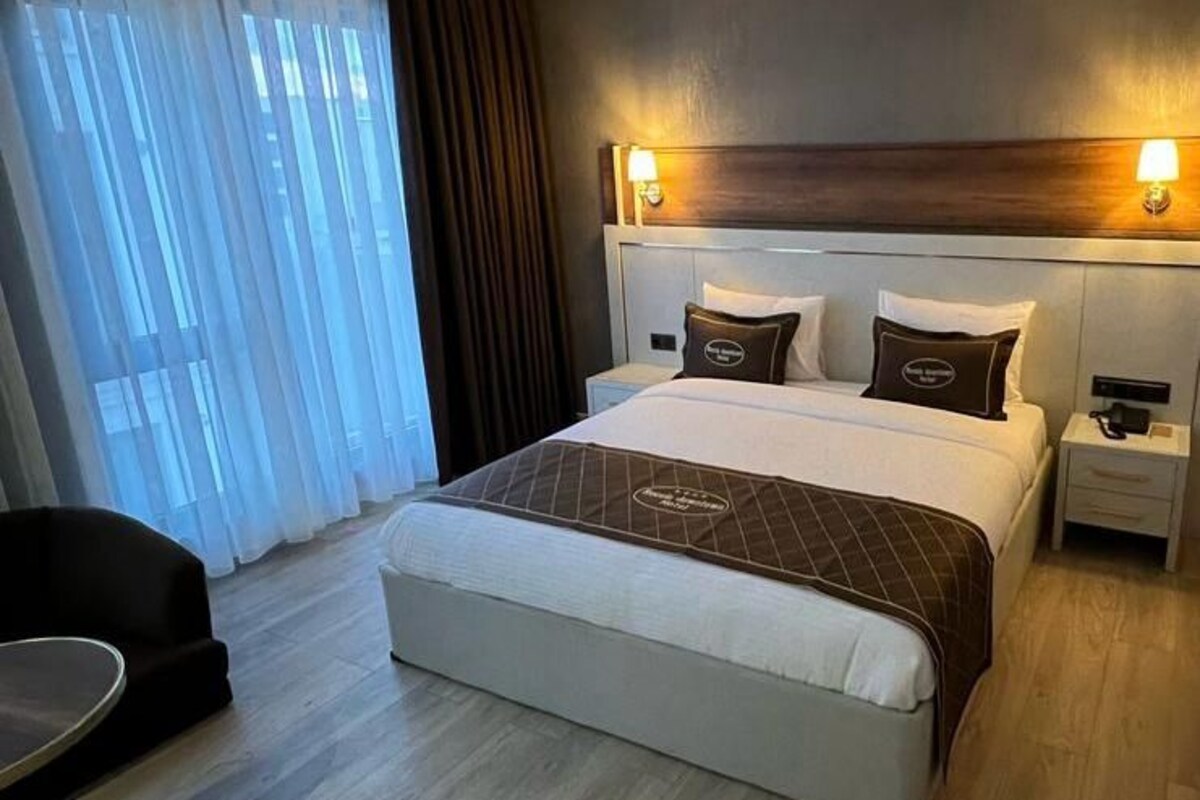 Quarto de hotel moderno com uma cama grande, mesas de cabeceira, uma escrivaninha e vista para a cidade através de grandes janelas.