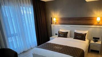 Una habitación de hotel moderna con una cama grande, mesitas de noche, un escritorio y vistas a la ciudad a través de amplios ventanales.