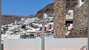 Une piscine dotée d’une plage en pierre, d’un escalier métallique et offrant une vue sur une ville côtière aux bâtiments blancs, avec une colline rocheuse en arrière-plan.
