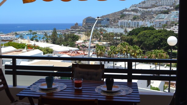 Un balcon avec une table et des chaises, donnant sur une ville côtière avec ses bâtiments, une plage et un ciel bleu dégagé.
