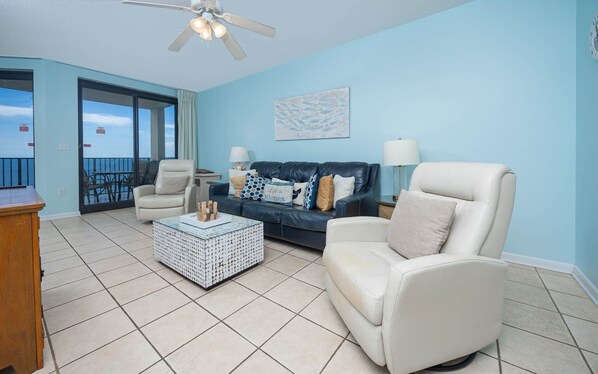 A living room with a blue wall, a ceiling fan, a white leather sofa, a blue sofa, and a coffee table with a decorative centerpiece. - Phoenix X 1405 1 Bedroom Condo (Orange Beach)