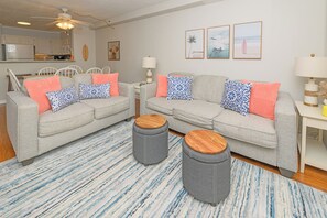 A living room with two grey sofas, wooden ottomans, a patterned rug, and a dining area with white chairs and a table. - Oceanfront 2 BR Condo with a Shared Pool! Beach House 302 (Garden City)